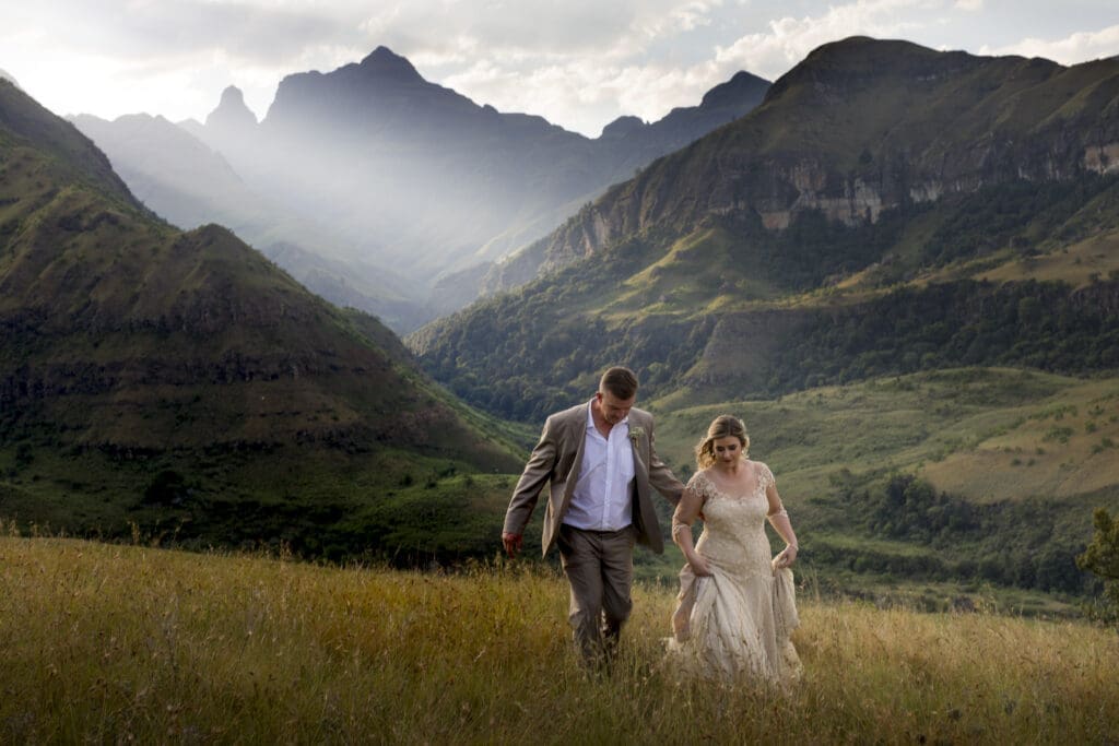 Newlyweds walking through long grass with beautiful mountain backdrop in Drakensberg