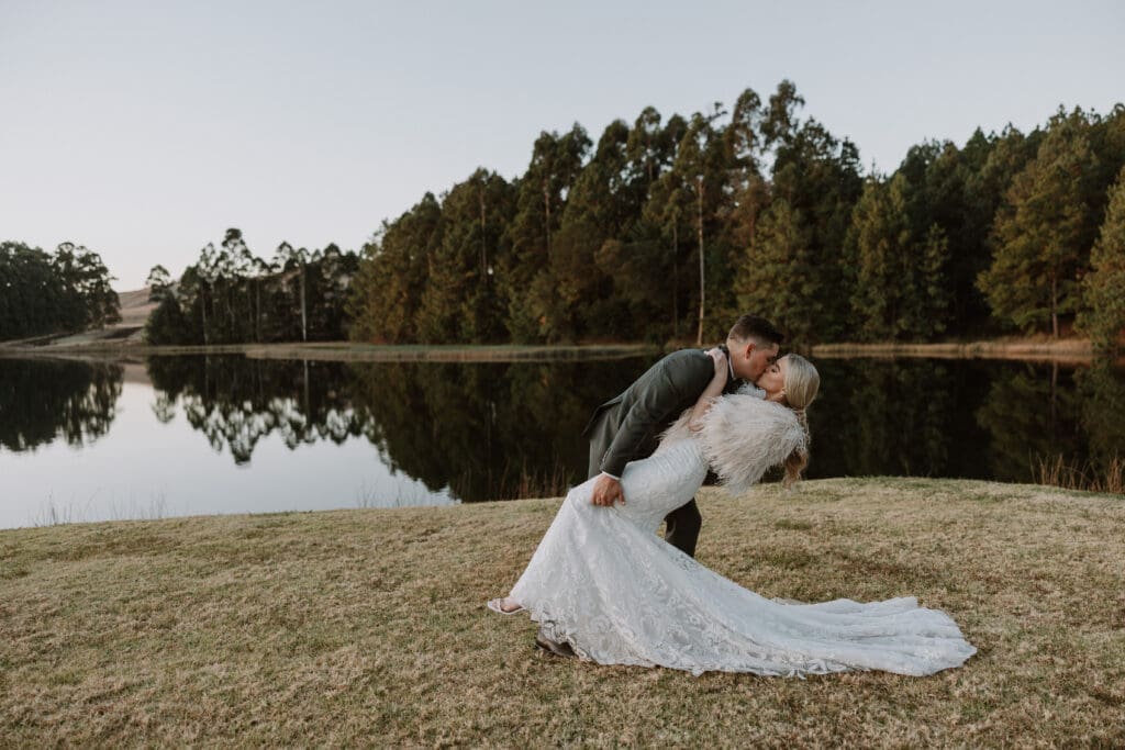 Groom dipping bride for a kiss in front of the water. Beautiful reflection in the water with trees on the other side.