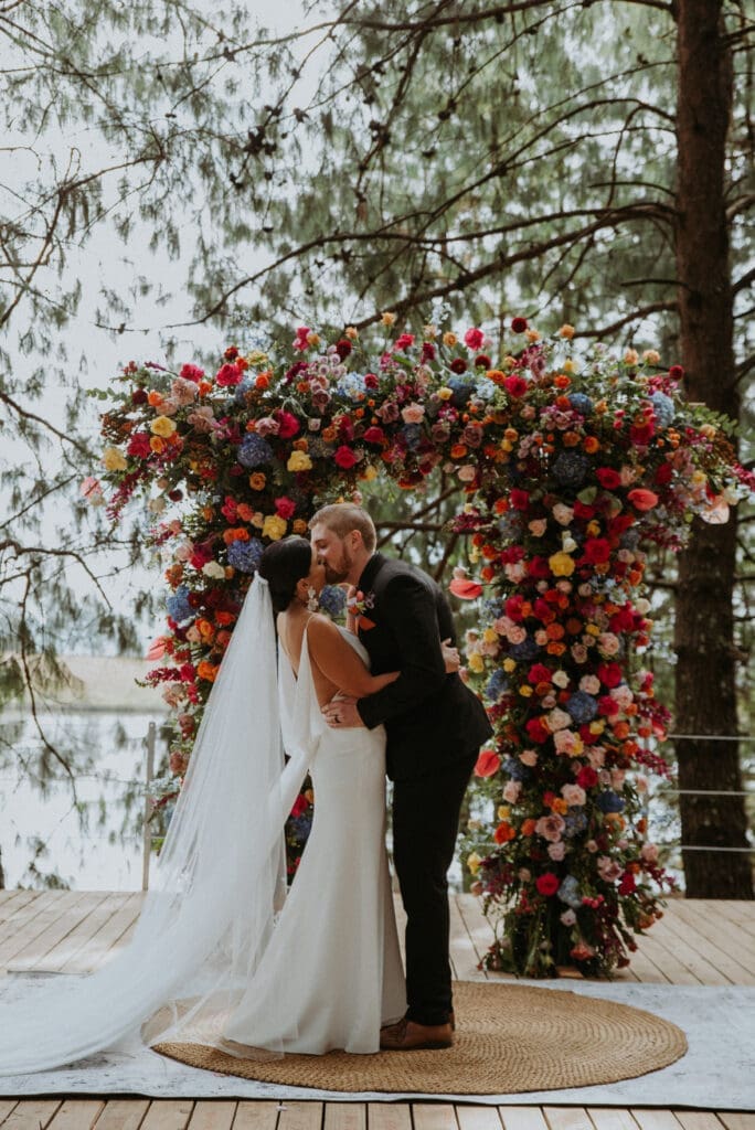Newlyweds exchanging first kiss in front of vibrant floral arch in forest setting
