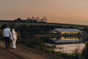 View of bride and groom walking back towards venue from the entrance road at dusk with reflections in the dam.