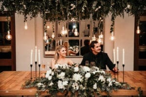Bride and groom sitting at dinner table with white floral arrangement in front.