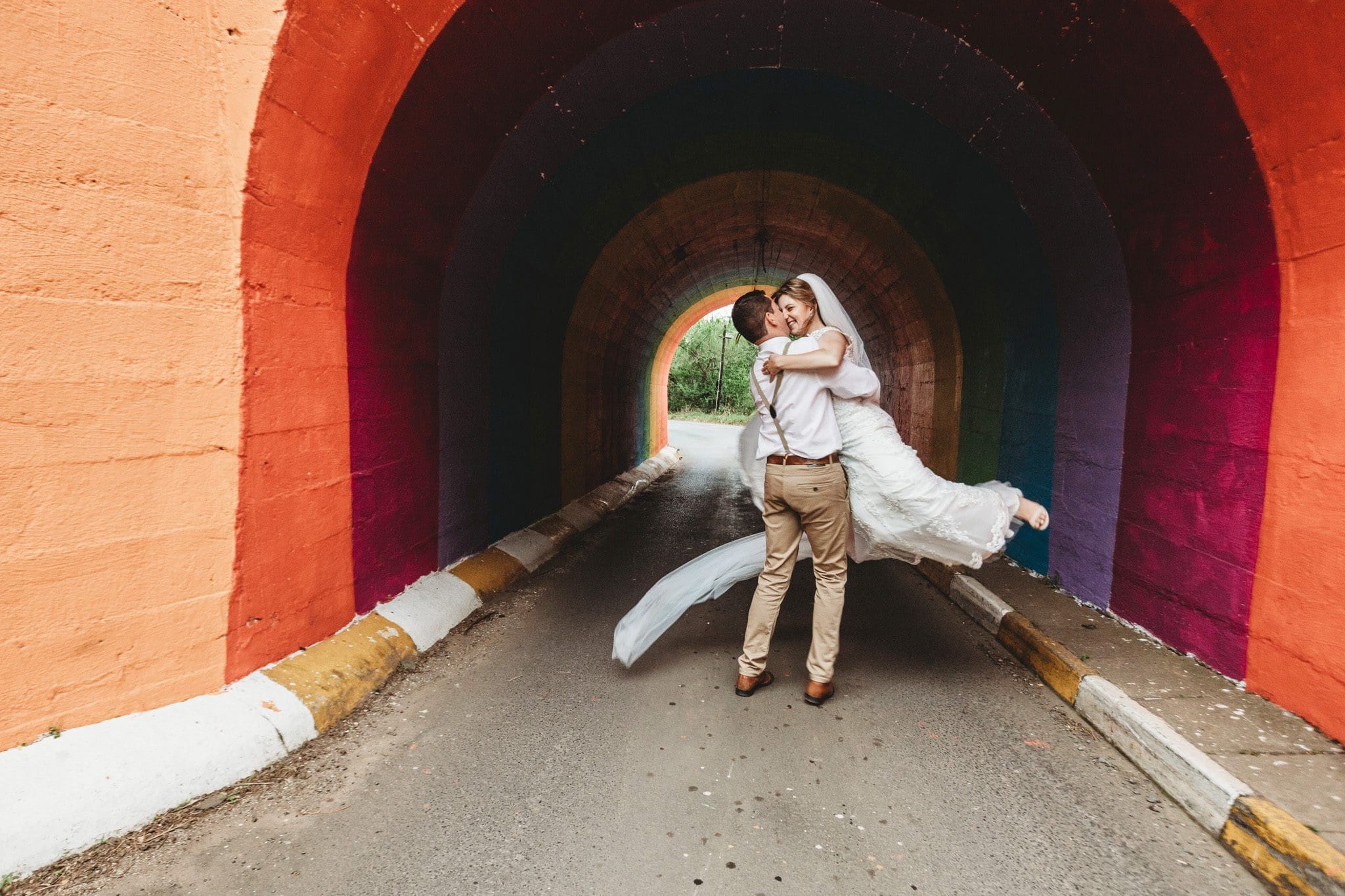 Groom lifts bride and swings her in front of colourful bridge
