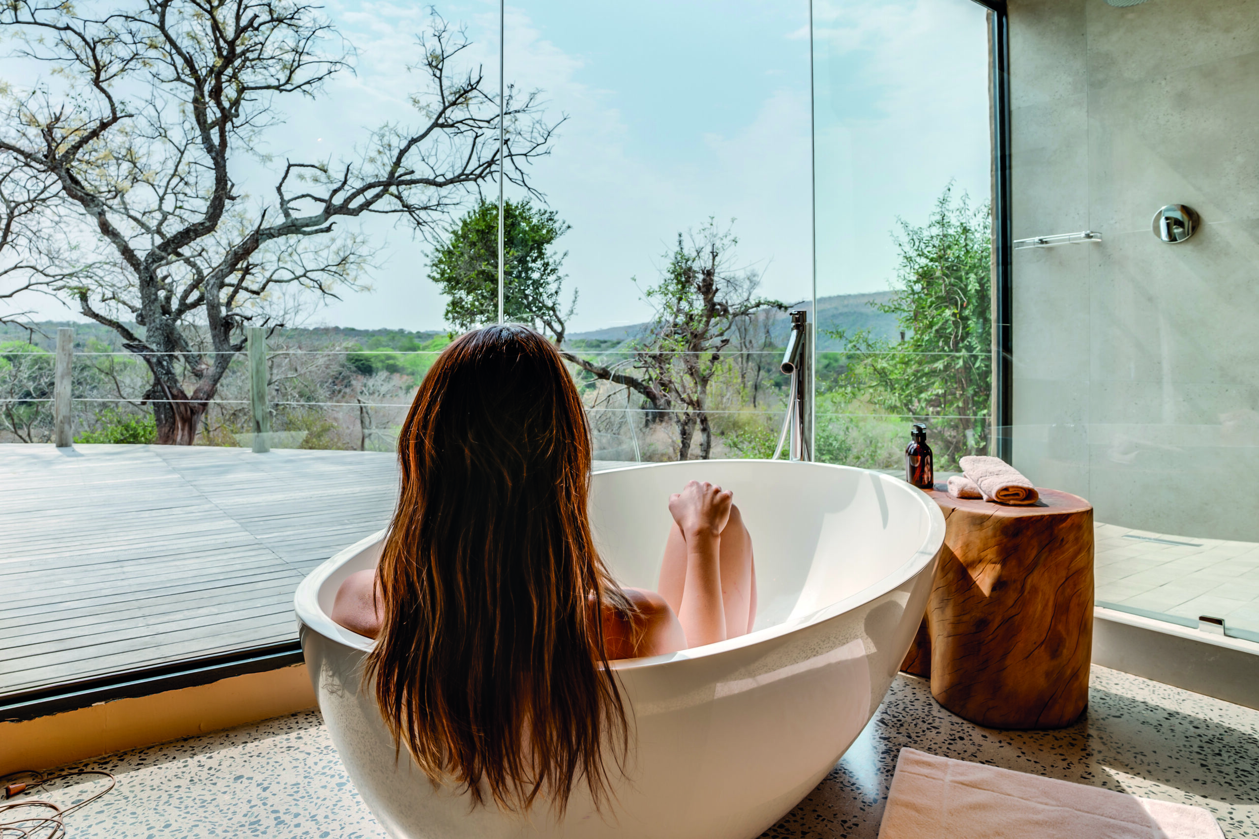 Woman taking a bath in glass enclosed bathroom overlooking African bush