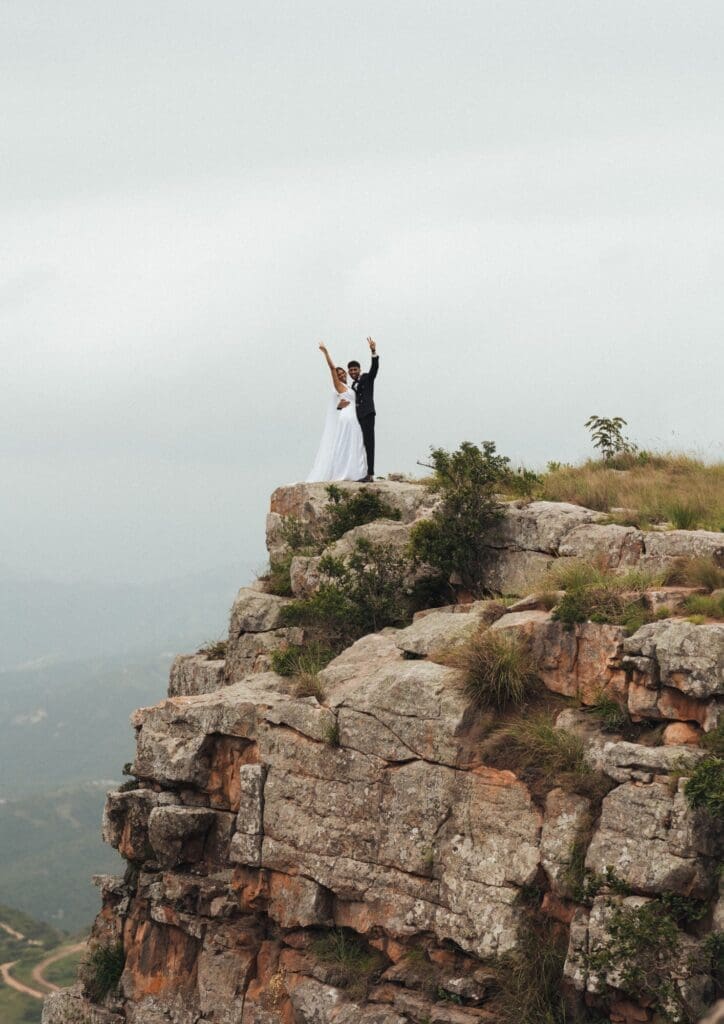 Bride and groom standing on cliff edge with arms in the air