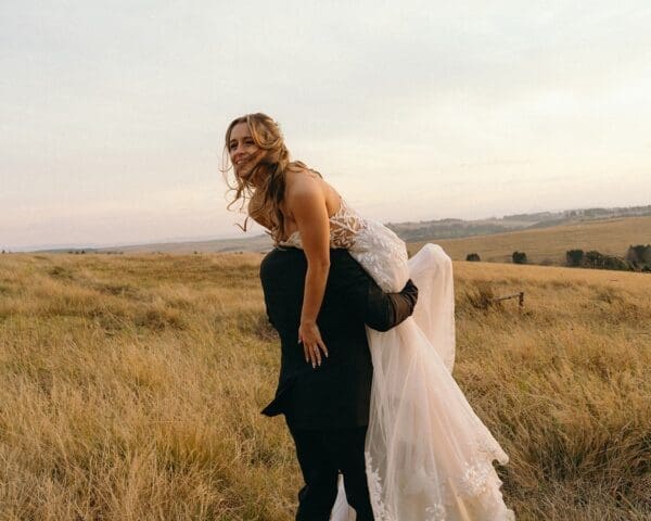 Bride being lifted onto grooms shoulder in open grass field