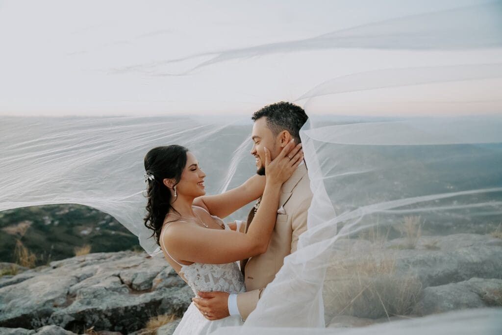 Bride and groom standing atop rocky ledge gazing at each other under wispy veil