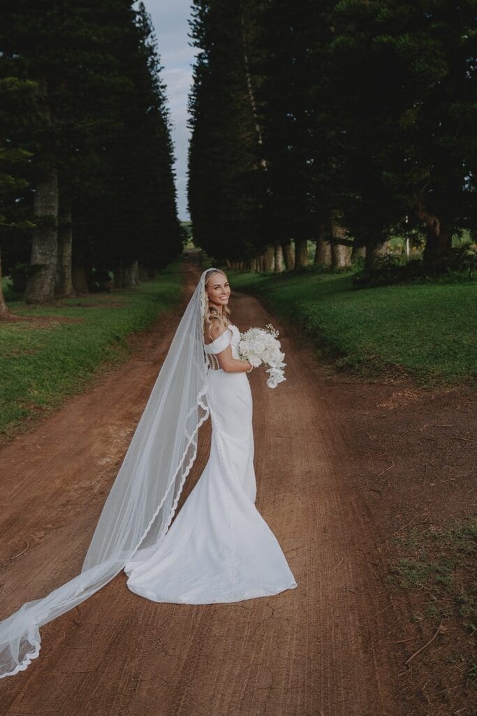 Bride standing on dirt road in avenue of trees looking back at the camera