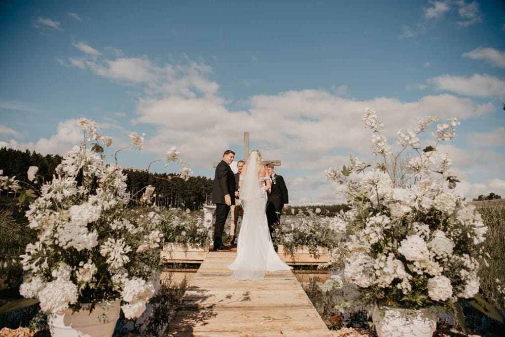 Couple exchanging vows at end of wooden deck with blue sky backdrop and white flowers