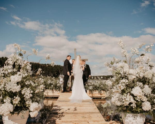 Couple exchanging vows at end of wooden deck with blue sky backdrop and white flowers