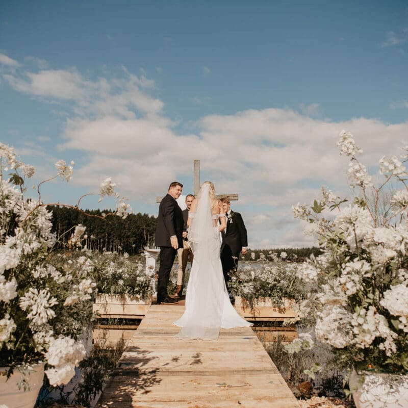 Couple exchanging vows at end of wooden deck with blue sky backdrop and white flowers