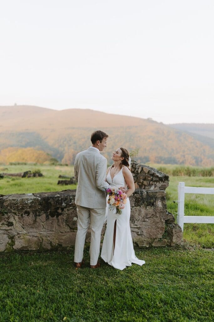 Bride and groom standing in grass field by low stone wall gazing into each others eyes