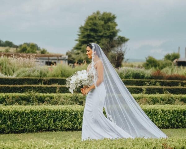Bride walking through low green hedges with trailing veil