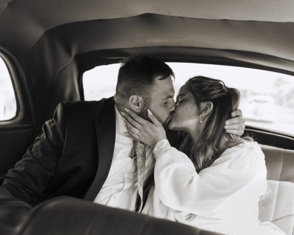 Black and white photo and video image of bride and groom kissing in the back of a vintage car.