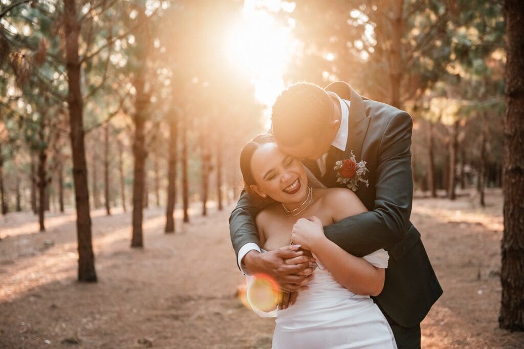 Groom embracing bride from behind as sun glows behind