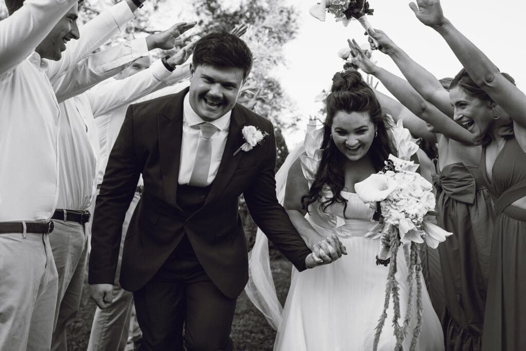 Black and white image of bride and groom walking through tunnel made by bridal party