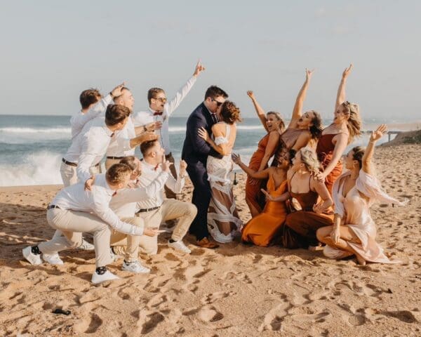 Bridal party celebrates as bride and groom kiss on the beach at Baron Beach House