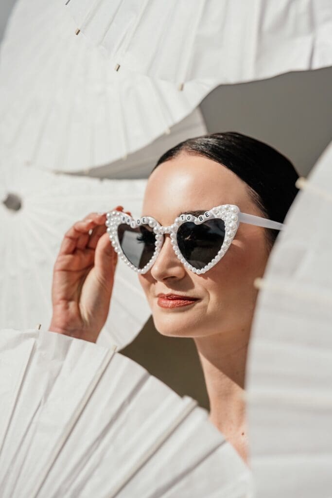 Close up of bride's face surrounded by white fans and wearing white heart shaped sunglasses