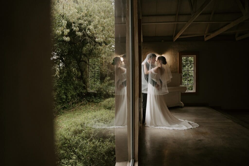 Bride and groom standing in chapel under bride's veil with reflection in the glass window