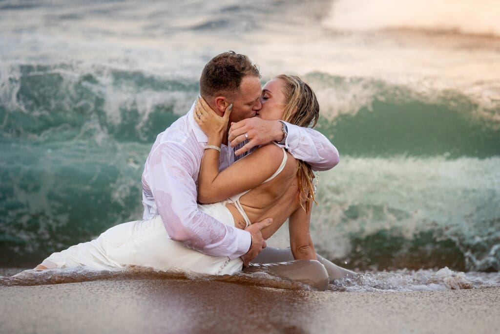 Bride and groom sitting in the waves at the beach sharing intimate kiss