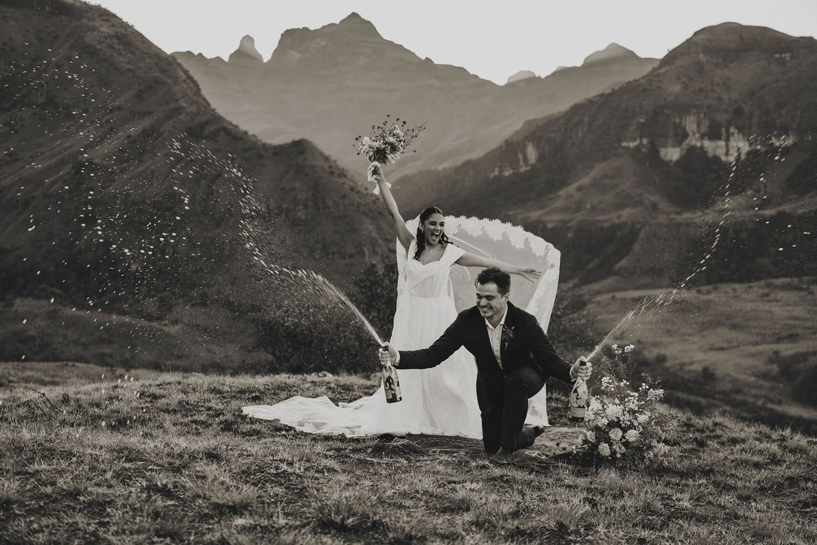 Black and white image of groom smashing champagne bottles on the floor for a dramatic spray in front of majestic mountain peaks and bride